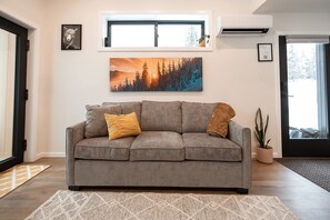 A living room with a grey sofa, two cushions, a painting above the sofa, and a potted plant. - Tinwood Cabins The Descent, Cozy 3 min to Rossland (Rossland)