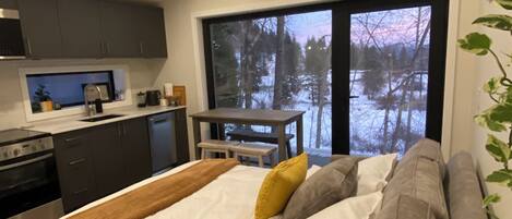 Een moderne slaapkamer met een groot bed, een zithoek en uitzicht op een besneeuwd bos door een groot raam.