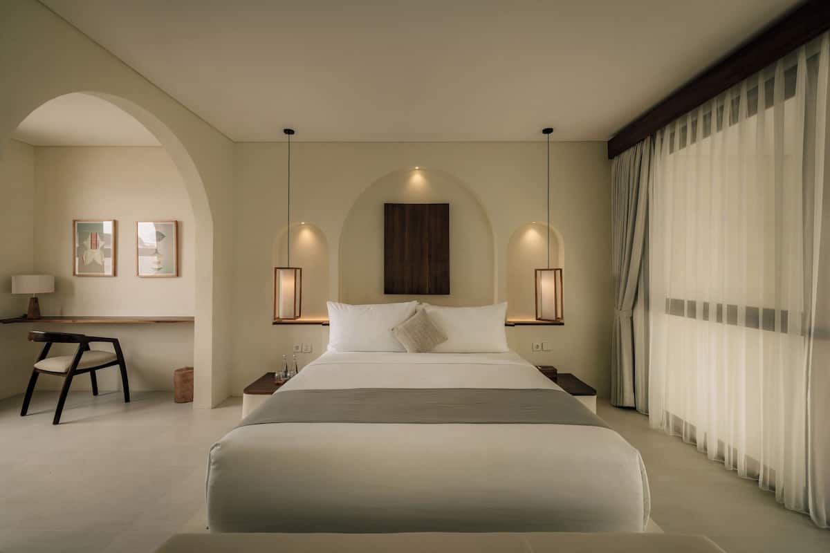 Une chambre d’hôtel avec un grand lit, deux tables de chevet, un bureau avec une chaise et une grande fenêtre avec des rideaux.