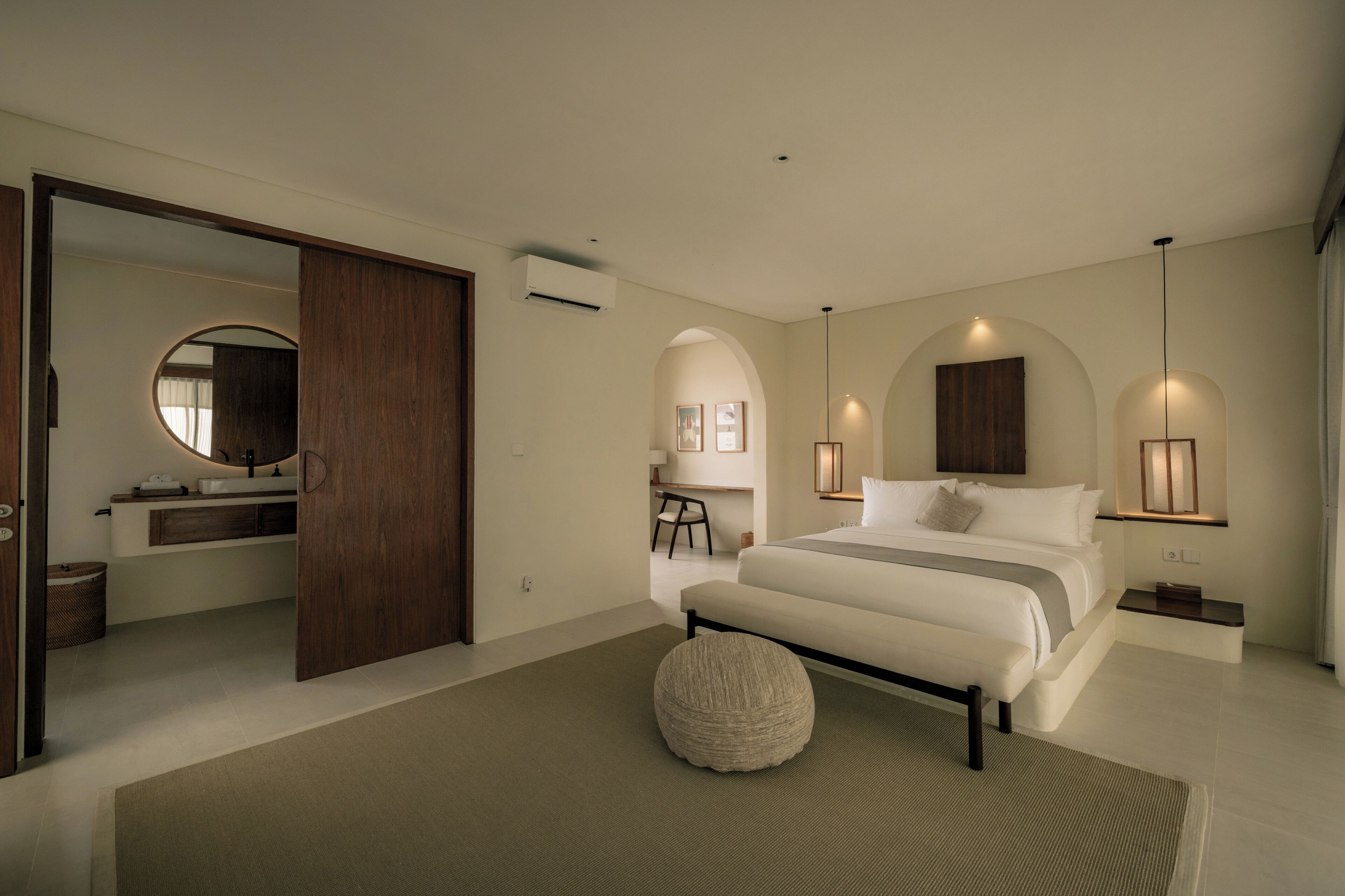 Un dormitorio moderno con una cama grande, un otoman redondo, un armario de madera y un baño con un espejo circular.