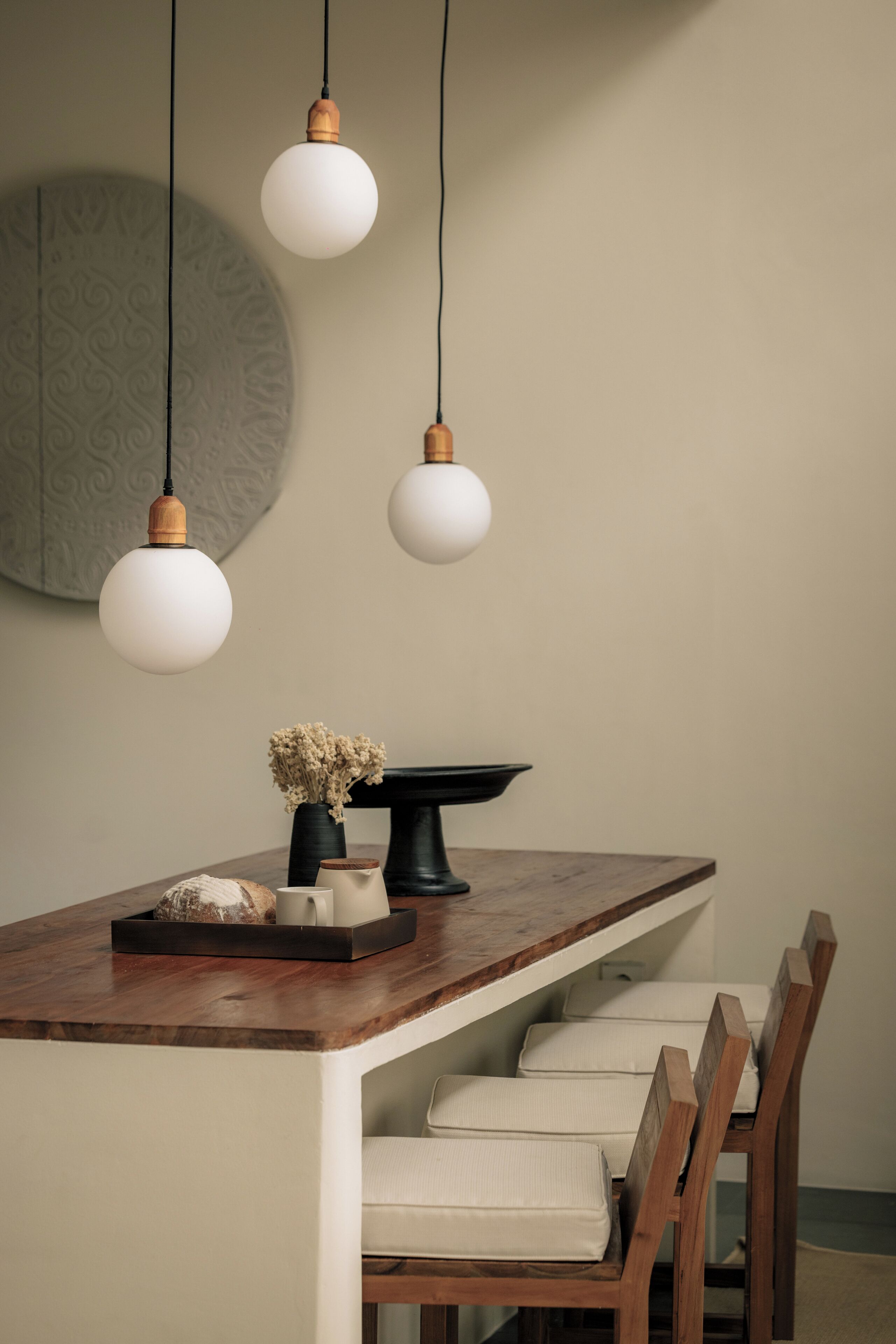 Un comedor con una mesa de madera, tres lámparas colgantes, una bandeja negra para servir y un florero con flores secas.
