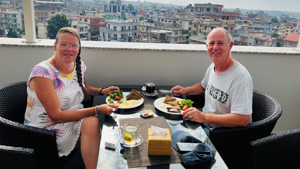 Dos personas cenando en una mesa con vistas a la ciudad, mostrando una combinación de edificios residenciales y una cadena montañosa distante.