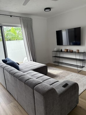 A modern living room with a grey sectional sofa, a flat-screen TV mounted on the wall, and a minimalist console table with decorative items. - 2Br - Walk to Beach and Dining (North Ward)
