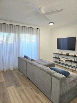 Un salon moderne avec un grand canapé d’angle gris, un téléviseur à écran plat fixé au mur et un ventilateur de plafond.