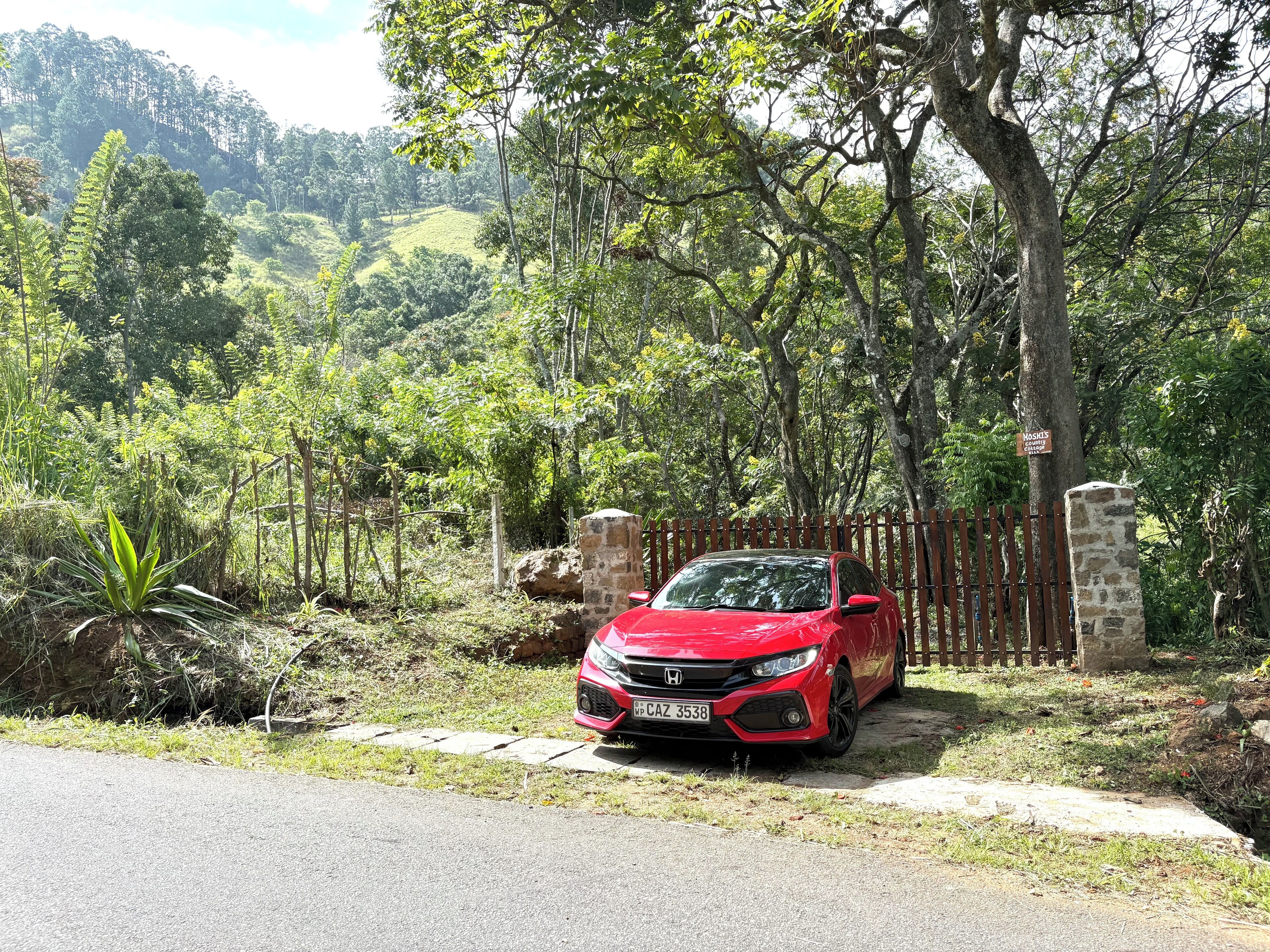 Une voiture rouge est garée sur un chemin pavé, entourée d’une végétation luxuriante et d’arbres.