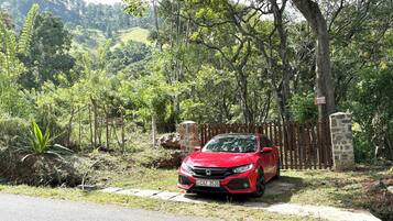 Une voiture rouge est garée sur un chemin pavé, entourée d’une végétation luxuriante et d’arbres.