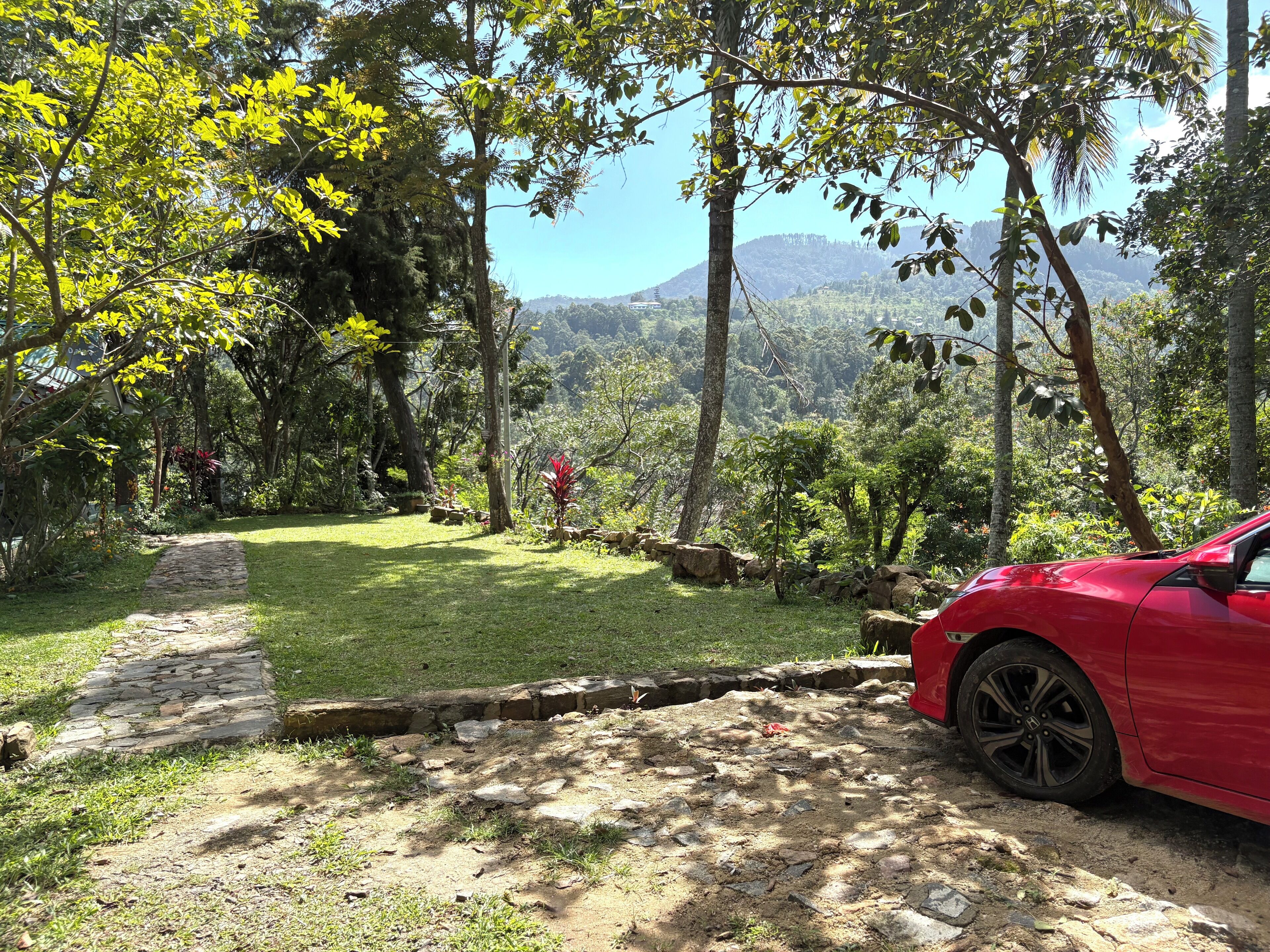 A red car parked on a dirt path with a stone wall and a grassy area with trees and a mountain in the background.