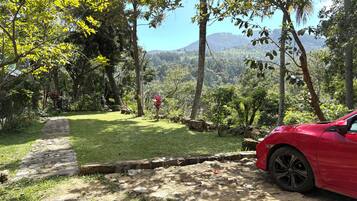A red car parked on a dirt path with a stone wall and a grassy area with trees and a mountain in the background.