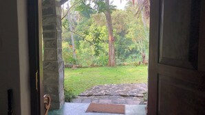 An open doorway leading to a stone-paved area with a doormat, surrounded by greenery and a stone wall. - Koshi's Lotus room Ella (Bandarawela)