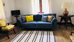 A living room with a blue sofa, yellow and patterned cushions, a striped rug, a wooden chair, a small table with a lamp, and a television on a stand. - Koshi's Lotus room Ella (Bandarawela)