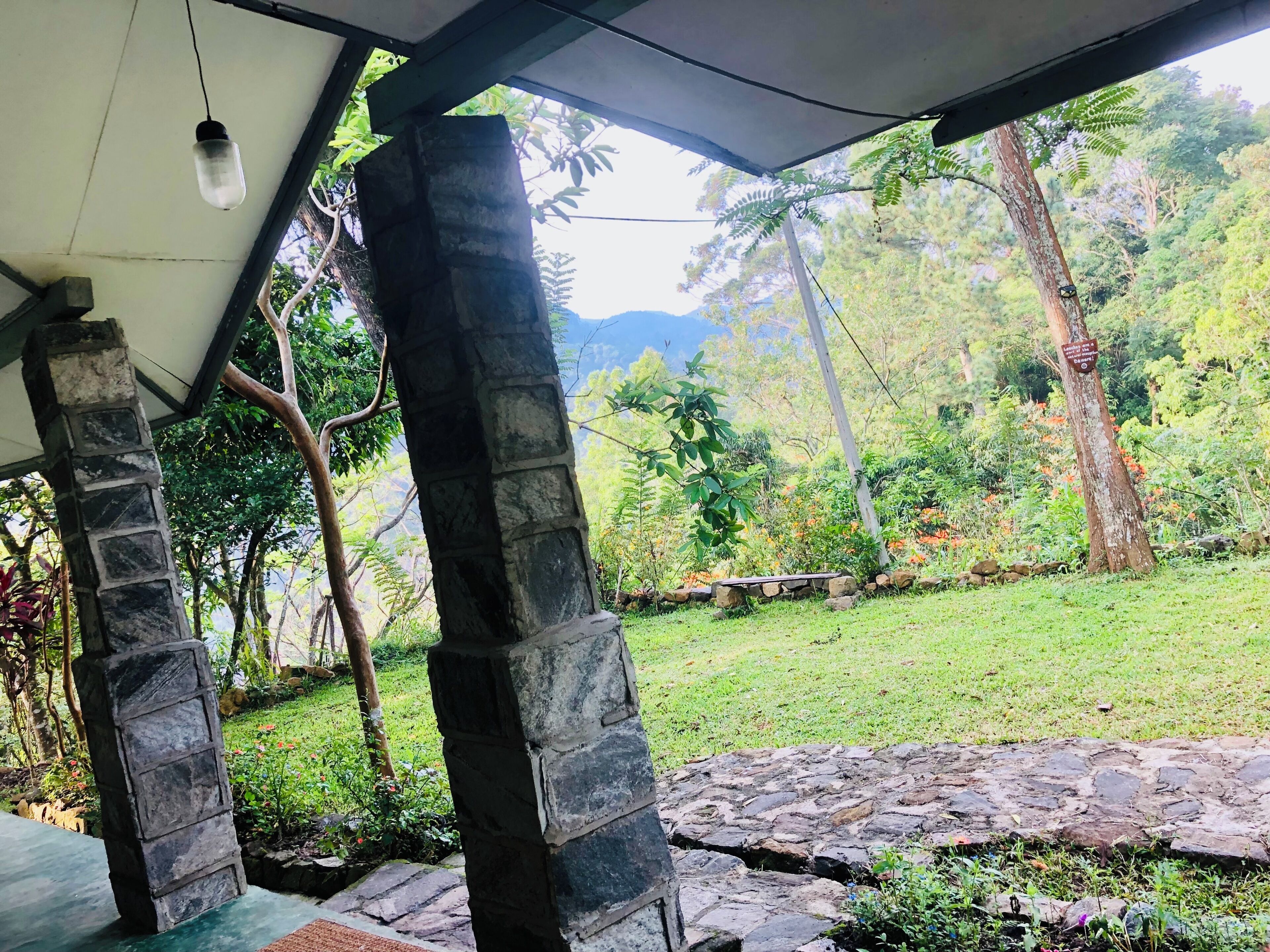 A covered outdoor area with stone pillars, a stone pathway, and a view of a lush garden with trees and a mountain in the background.
