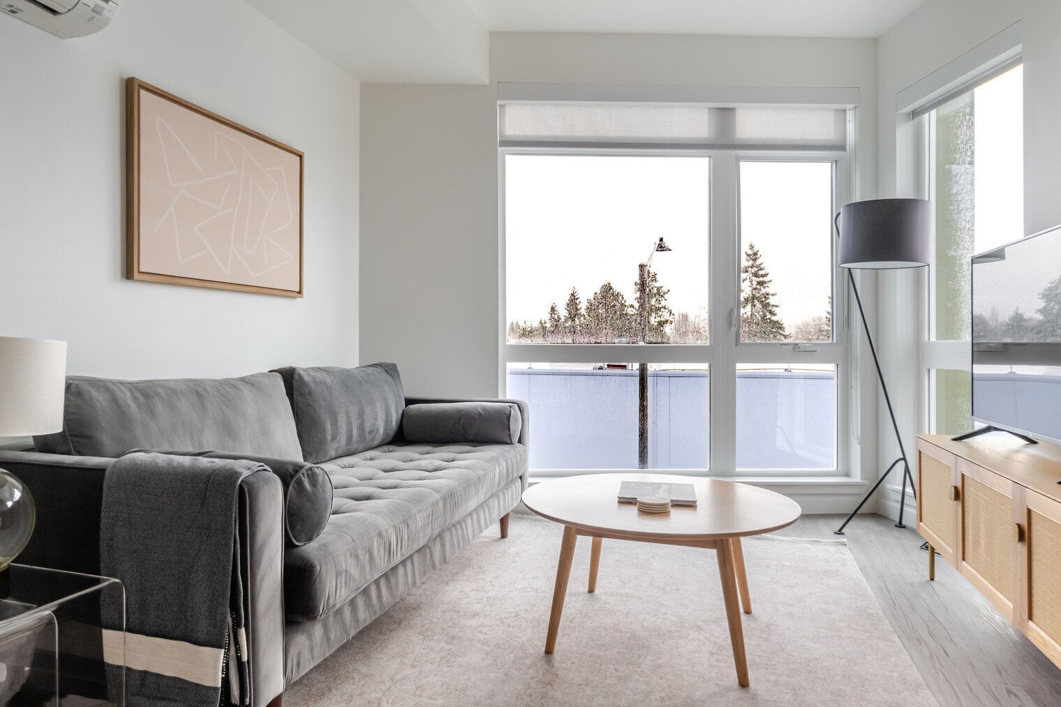 A modern living room with a grey sofa, a round coffee table, and a large window with a view of trees and a building.