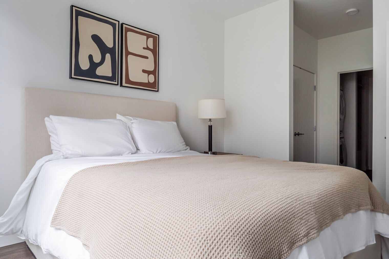 Un lit bien fait, avec du linge de lit blanc et une couverture beige, une lampe de chevet et deux tableaux abstraits accrochés au mur.