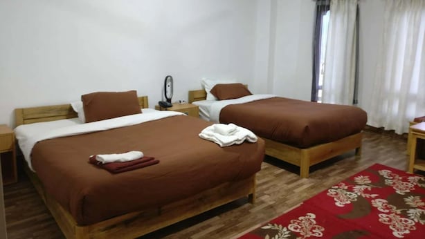 Une chambre d’hôtel avec deux lits simples, des têtes de lit en bois, des murs blancs, un plancher en bois, un tapis rouge à motifs et une petite table avec une chaise.
