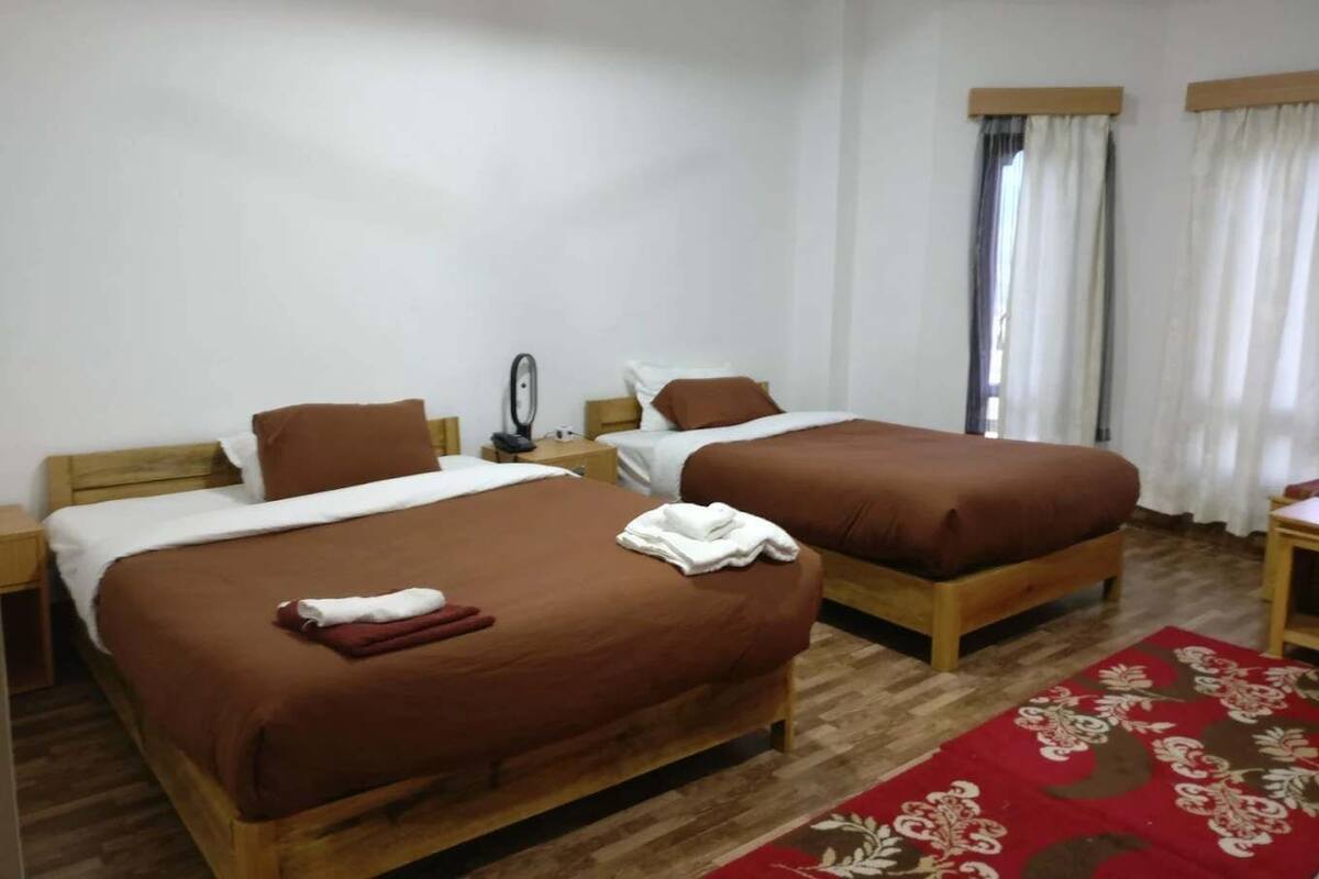 A hotel room with two single beds, wooden headboards, white walls, a wooden floor, a red patterned rug, and a small table with a chair.