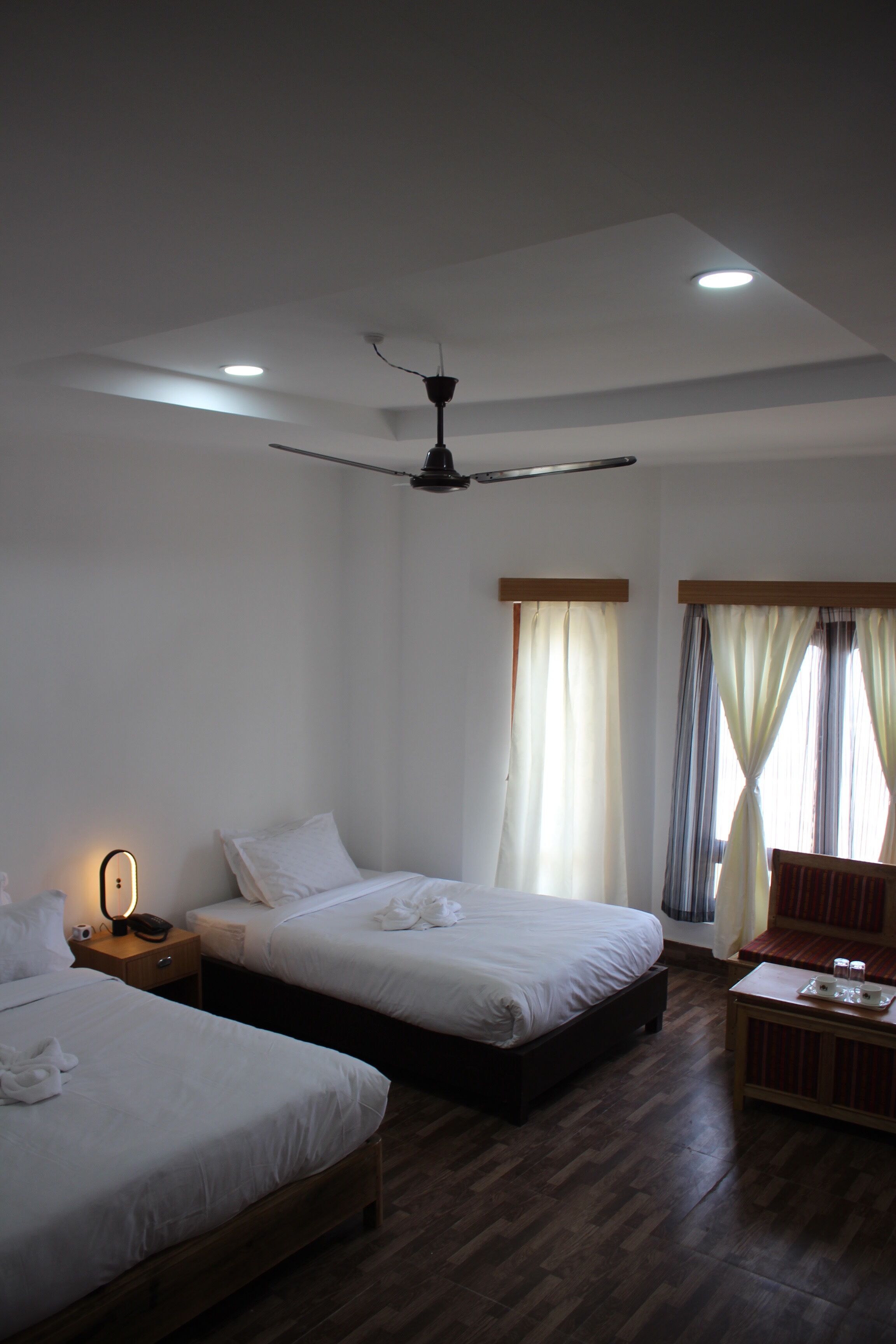 Une chambre d’hôtel avec deux lits, un ventilateur de plafond, une table de chevet avec une lampe, et un petit coin salon avec une table et des chaises.