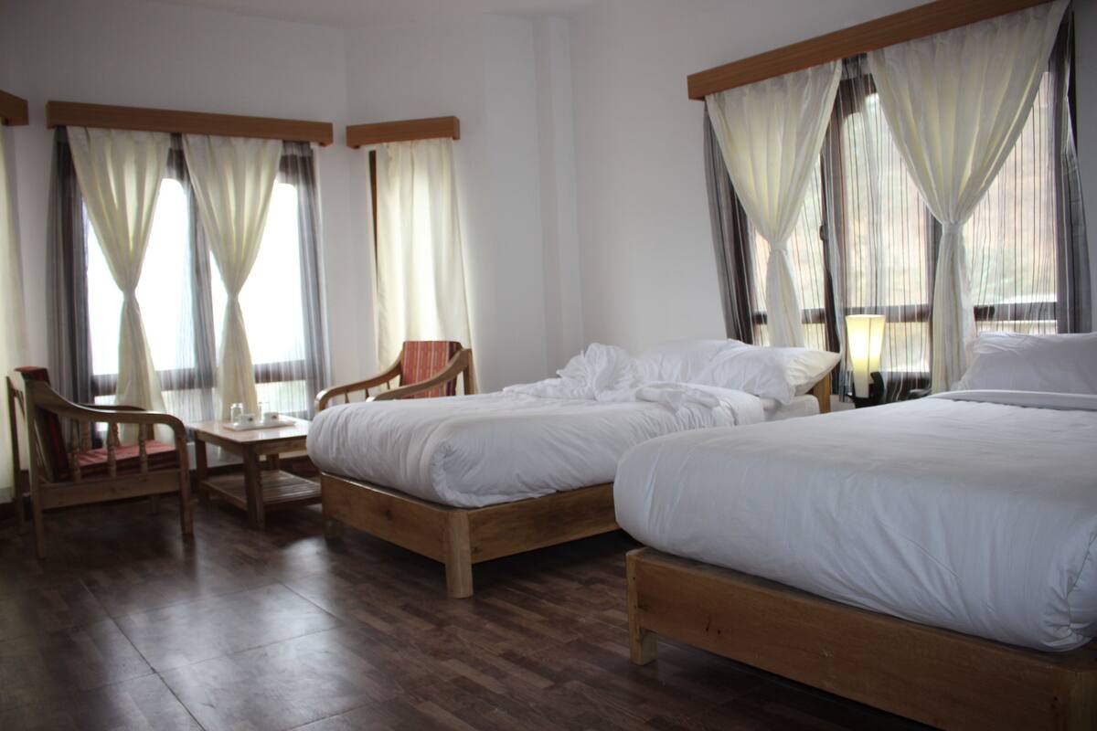 A hotel room with two wooden beds, a wooden chair, a small table, and sheer curtains.