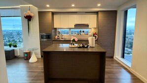 A modern kitchen with a central island, built-in appliances, and large windows offering a city view. - New 2 Bedroom Home Near Parks + Transit @skytrain &Plaza (Surrey)