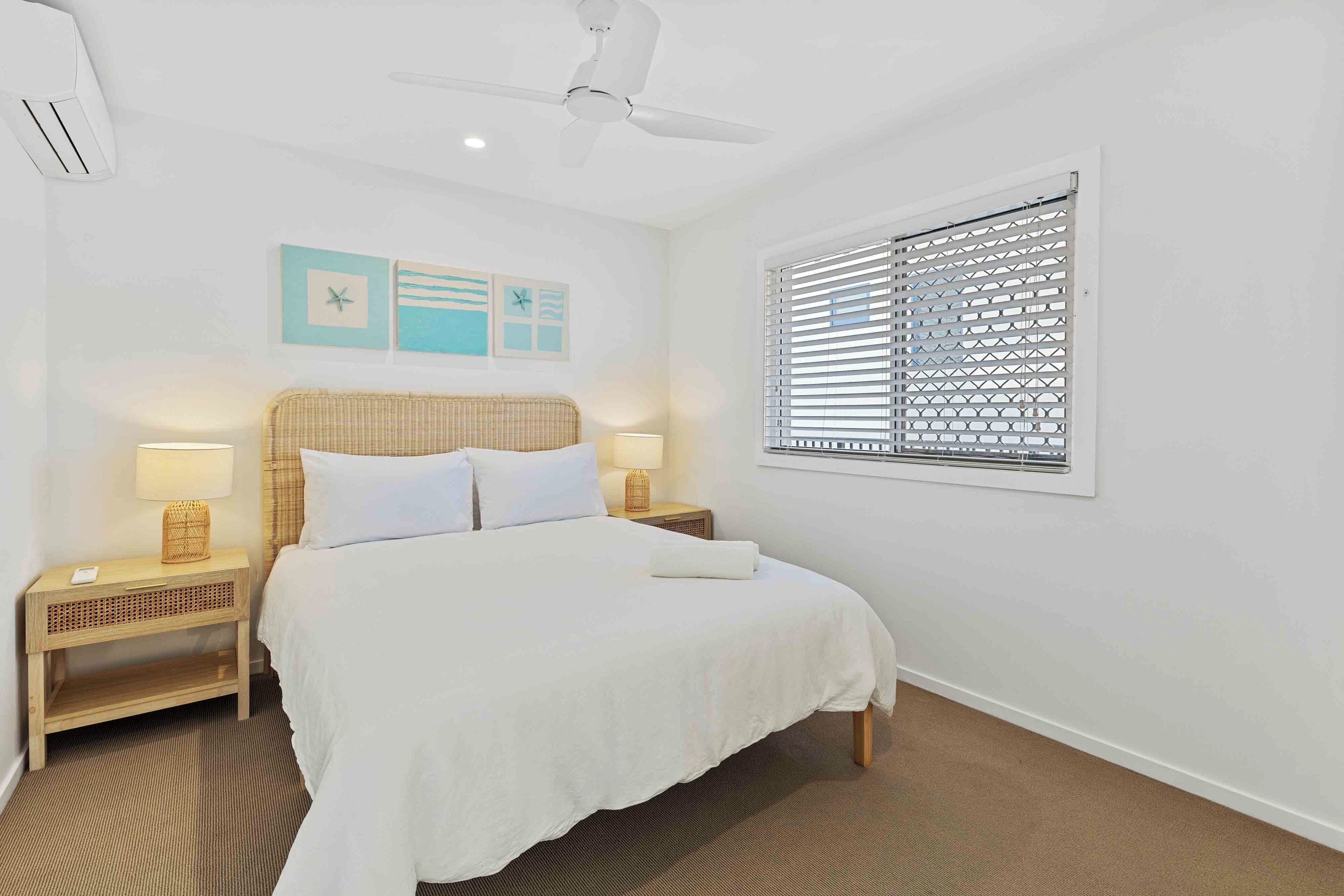 A hotel room with a bed, two bedside tables with lamps, a ceiling fan, a window with blinds, and wall art.
