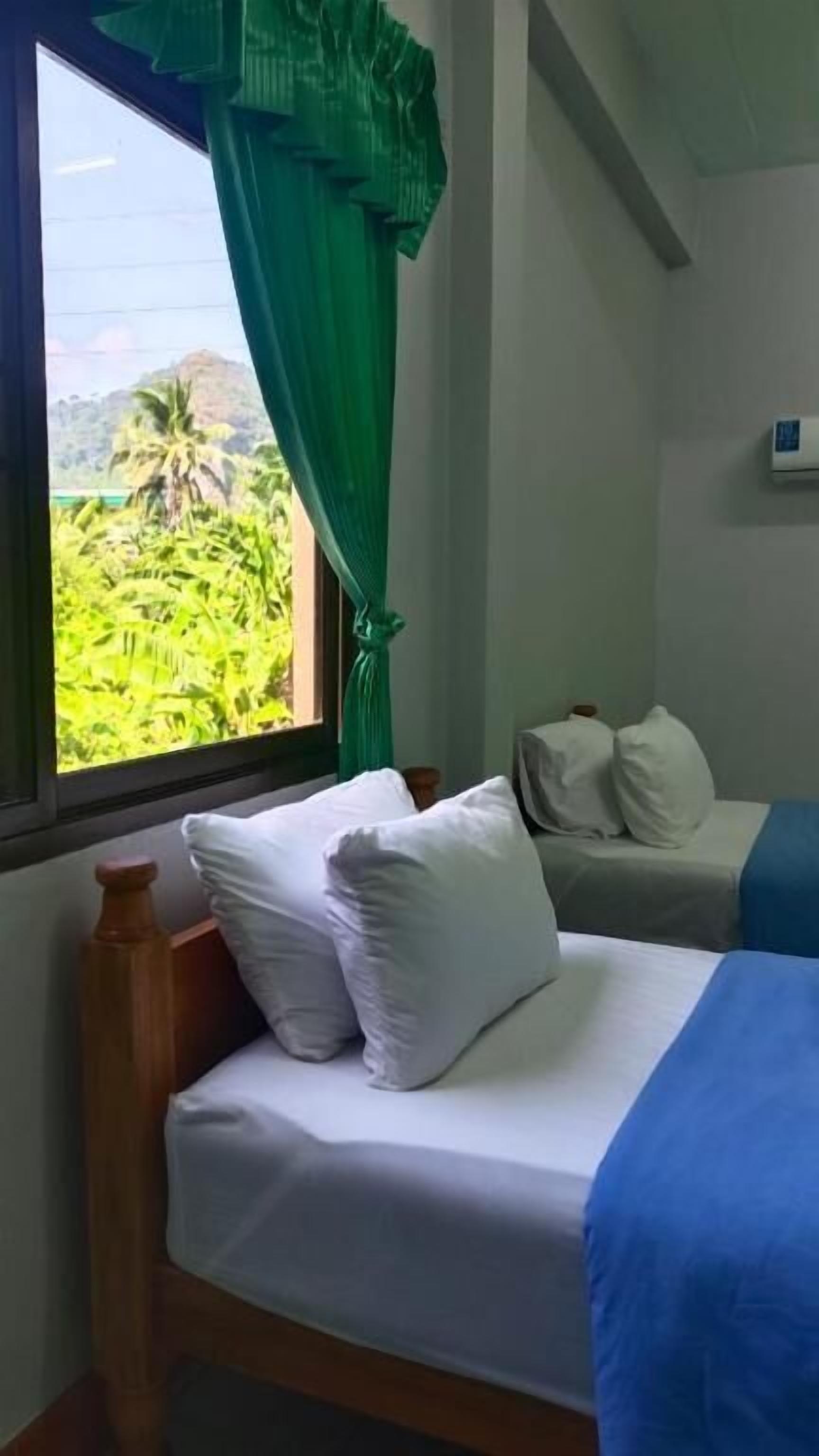 Ein Zimmer mit zwei Betten, eines mit weißer Bettwäsche und das andere mit blauer Bettwäsche. Ein Fenster bietet Blick auf grüne Vegetation.