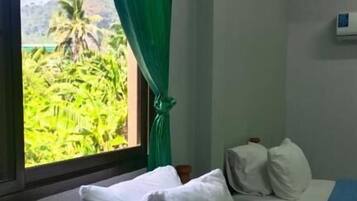 Ein Zimmer mit zwei Betten, eines mit weißer Bettwäsche und das andere mit blauer Bettwäsche. Ein Fenster bietet Blick auf grüne Vegetation.