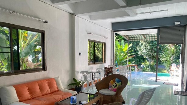 A modern living room with a sofa, wicker chairs, a glass coffee table, and a view of a pool and greenery. - Mountain View Hostel - Krabi Airport (Krabi)