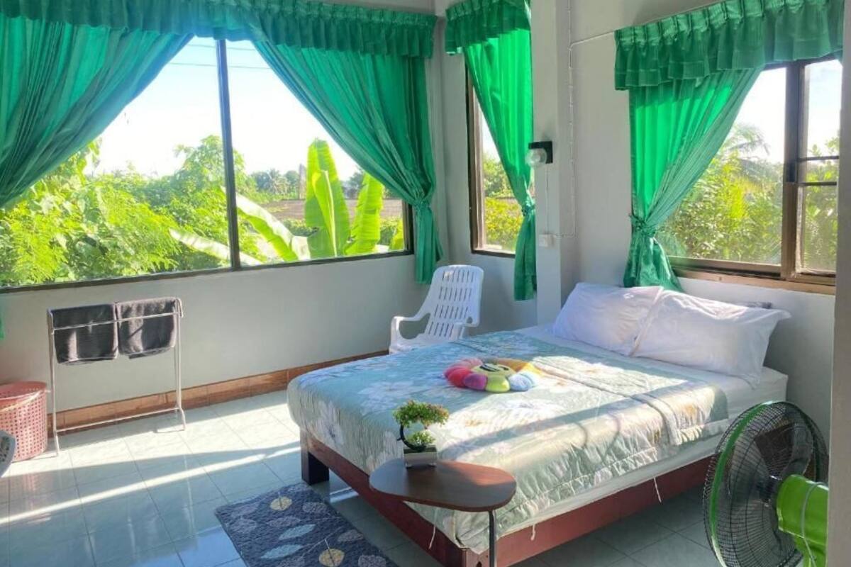 Une chambre avec un lit, une chaise, un ventilateur et une vue sur la verdure environnante.