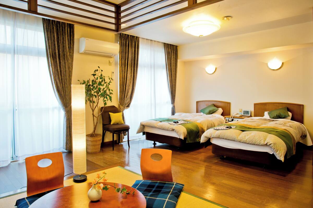 A hotel room with two beds, a dining table, a chair, a plant, and a window with curtains.
