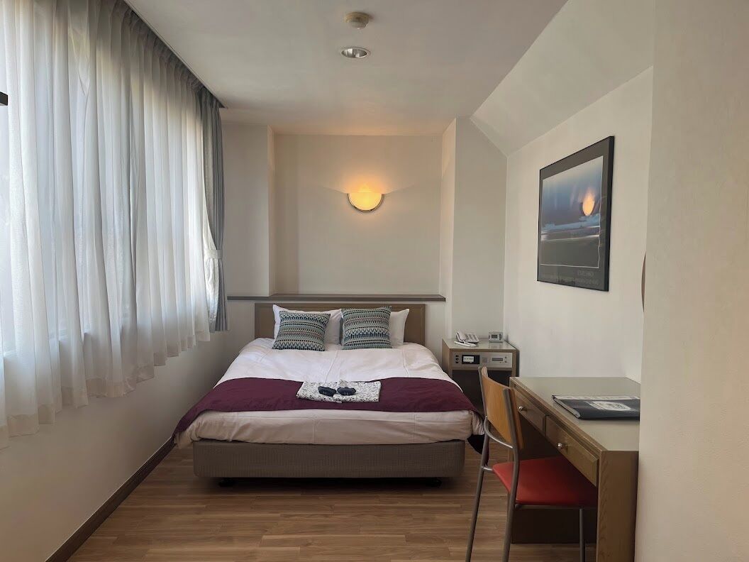 A hotel room with a bed, a desk, a chair, a nightstand, a wall-mounted lamp, and a painting.