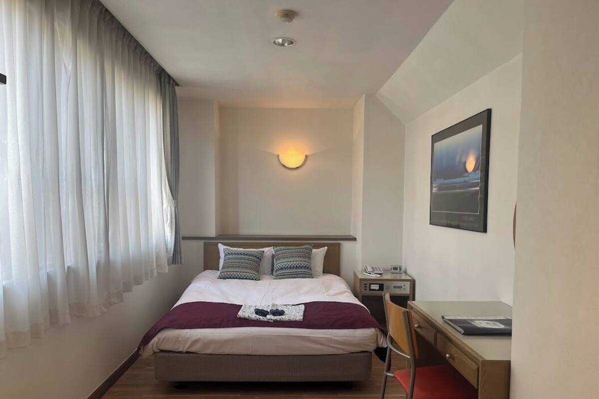 A hotel room with a bed, a desk, a chair, a nightstand, a wall-mounted lamp, and a painting.