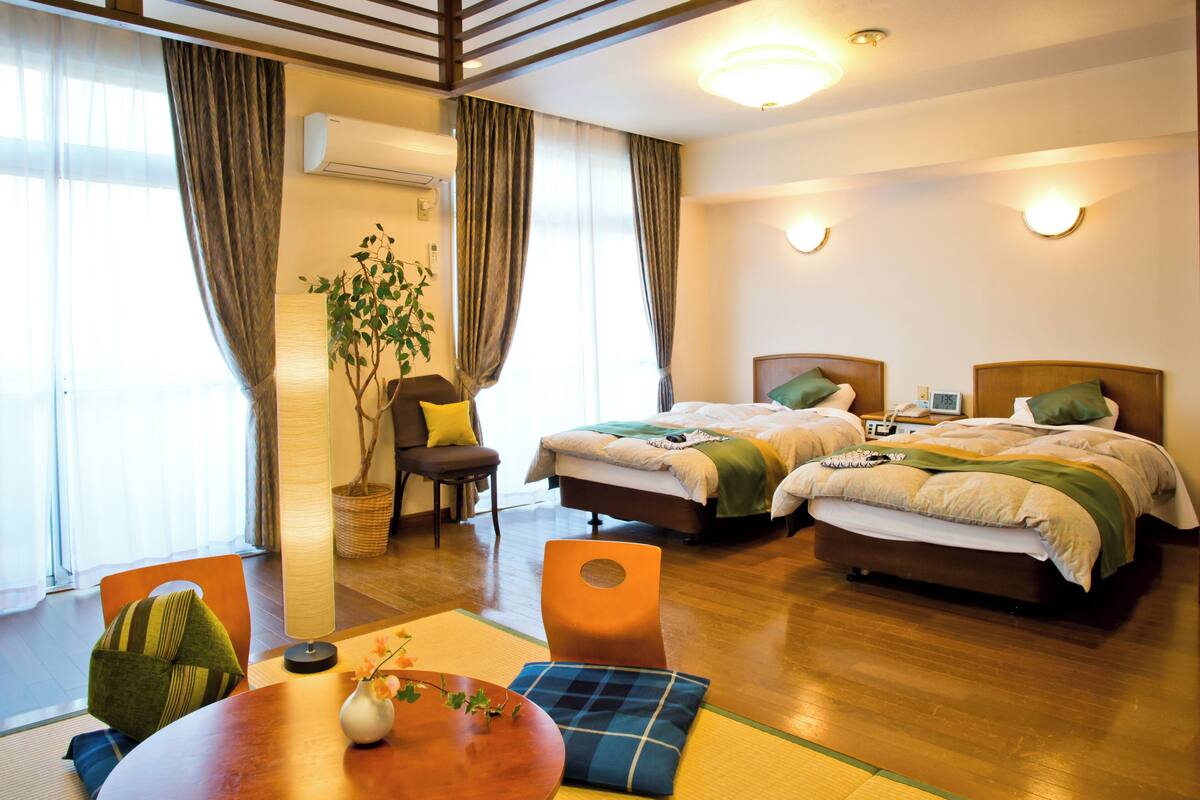 A hotel room with two beds, a dining table, a chair, a lamp, and a potted plant.