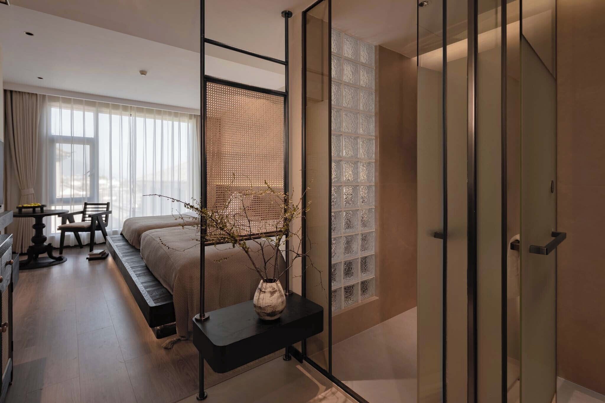 A modern bedroom with a bed, a desk, and a chair. There is a window with blinds and a decorative plant on a side table.