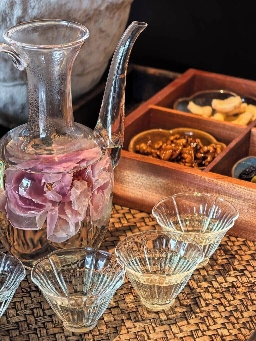 A clear glass pitcher filled with pink flowers, accompanied by three clear glass cups and a wooden box containing nuts and dried fruits.