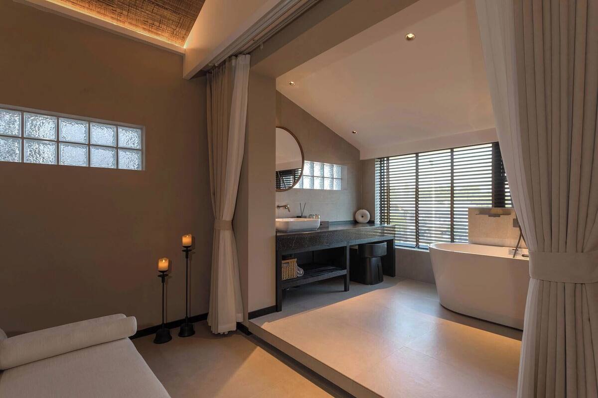 Een moderne badkamer met een groot ligbad, een wastafelmeubel met een wastafel en een spiegel. Er staan kaarsen op de vloer en er is een raam met zonwering.