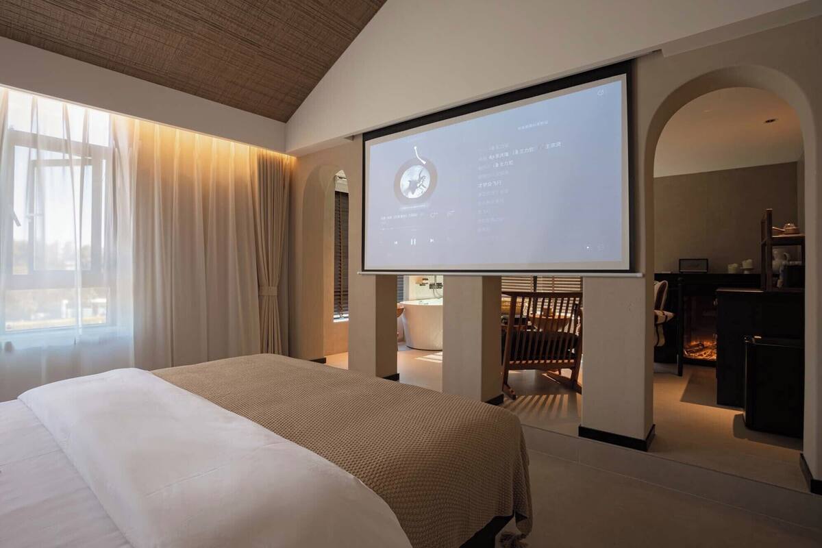 Een moderne hotelkamer met een groot bed, een televisiescherm waarop een menukaart staat en uitzicht door een groot raam.