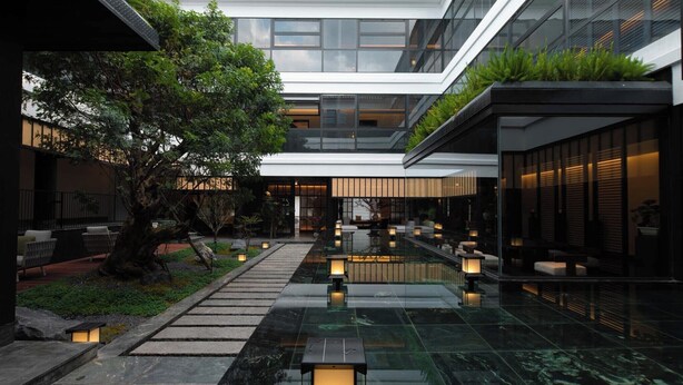 Un bâtiment moderne doté d’une façade en verre, d’un espace extérieur aménagé pour s’asseoir et d’un élément décoratif aquatique réfléchissant.