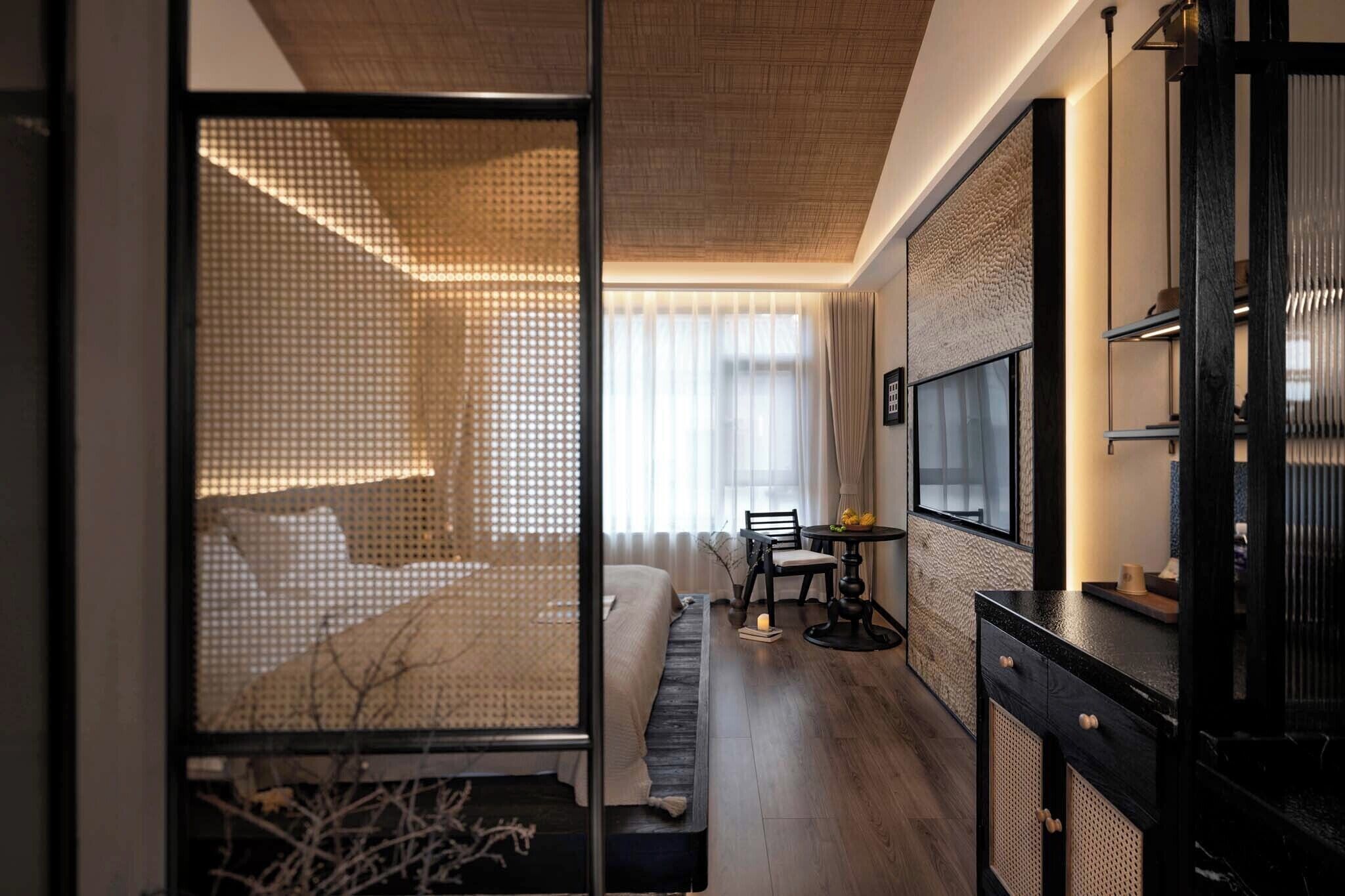Una habitación de hotel moderna con cama, una pequeña mesa con sillas, un televisor y un ventanal con cortinas translúcidas.