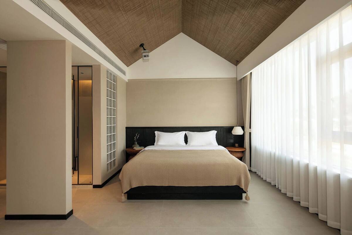 Een moderne slaapkamer met een groot bed, een nachtkastje, een lamp en een uitzicht op een stadsbeeld door grote ramen.
