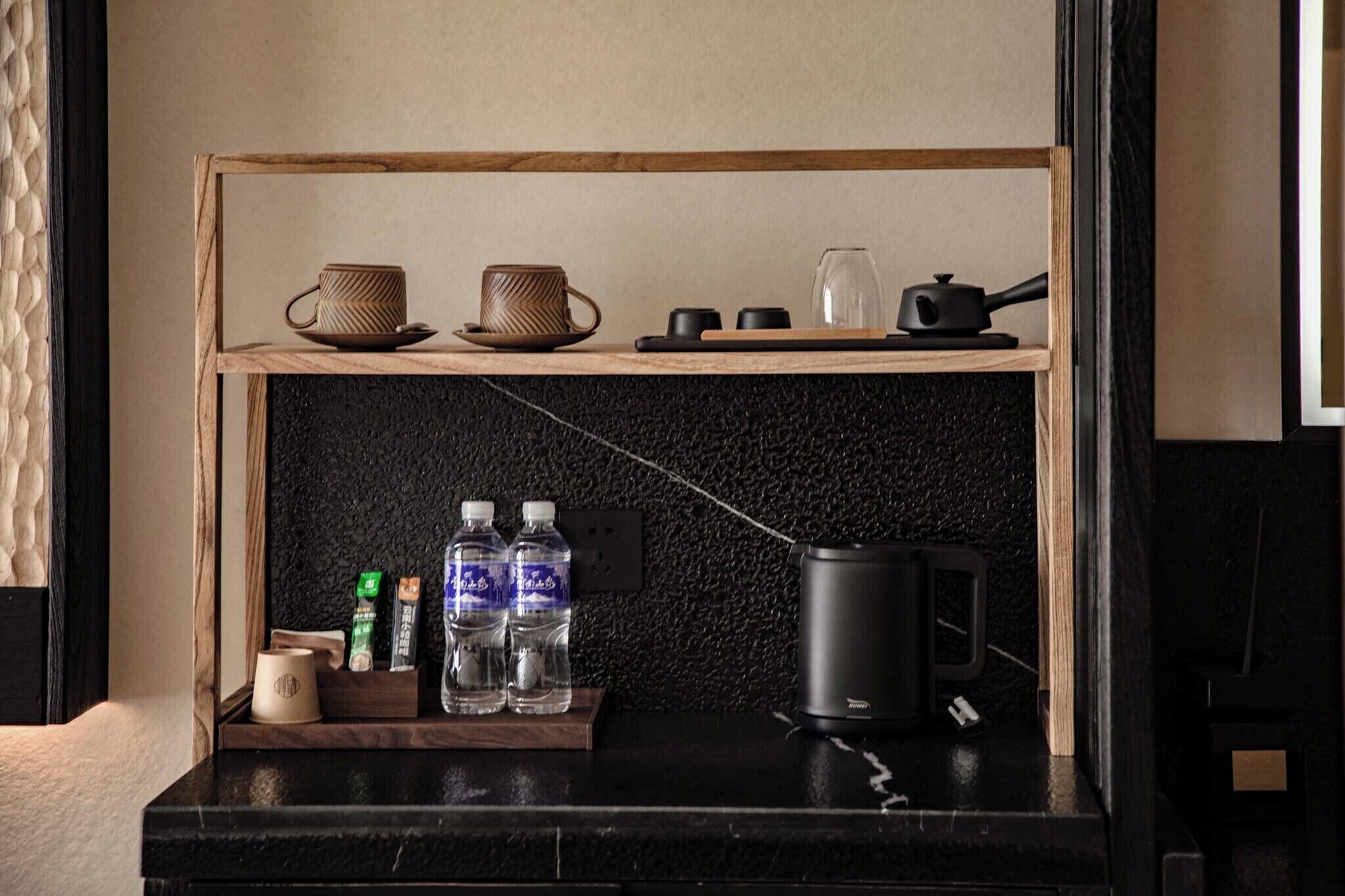 Una kitchenette con repisas de madera que sostiene dos tazas, un vaso y una tetera. Debajo de la repisa, hay botellas de agua y una cafetera.