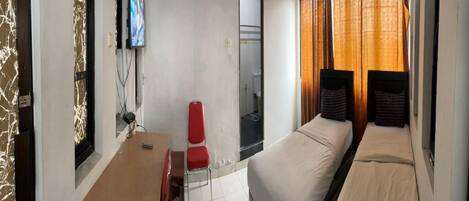 Une chambre d’hôtel de taille réduite, comprenant un lit simple, un petit bureau avec une chaise, un téléviseur fixé au mur et une fenêtre avec des rideaux.