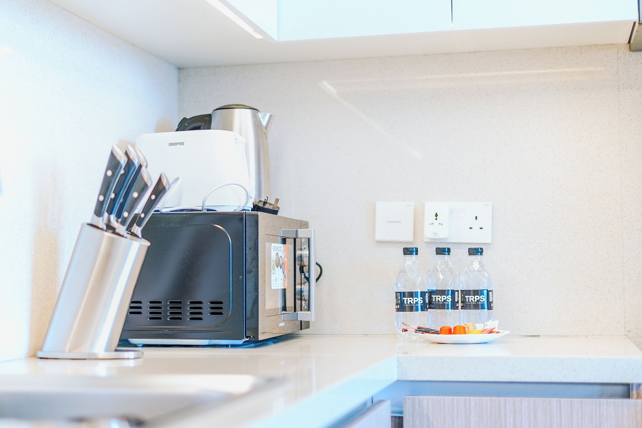 Una kitchenette con cafetera, bloque de cuchillos y dos botellas de agua TRP sobre la encimera.