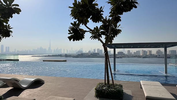 Uma área de piscina com vista para o horizonte da cidade através de um corpo d'água, uma pérgula e cadeiras de descanso.