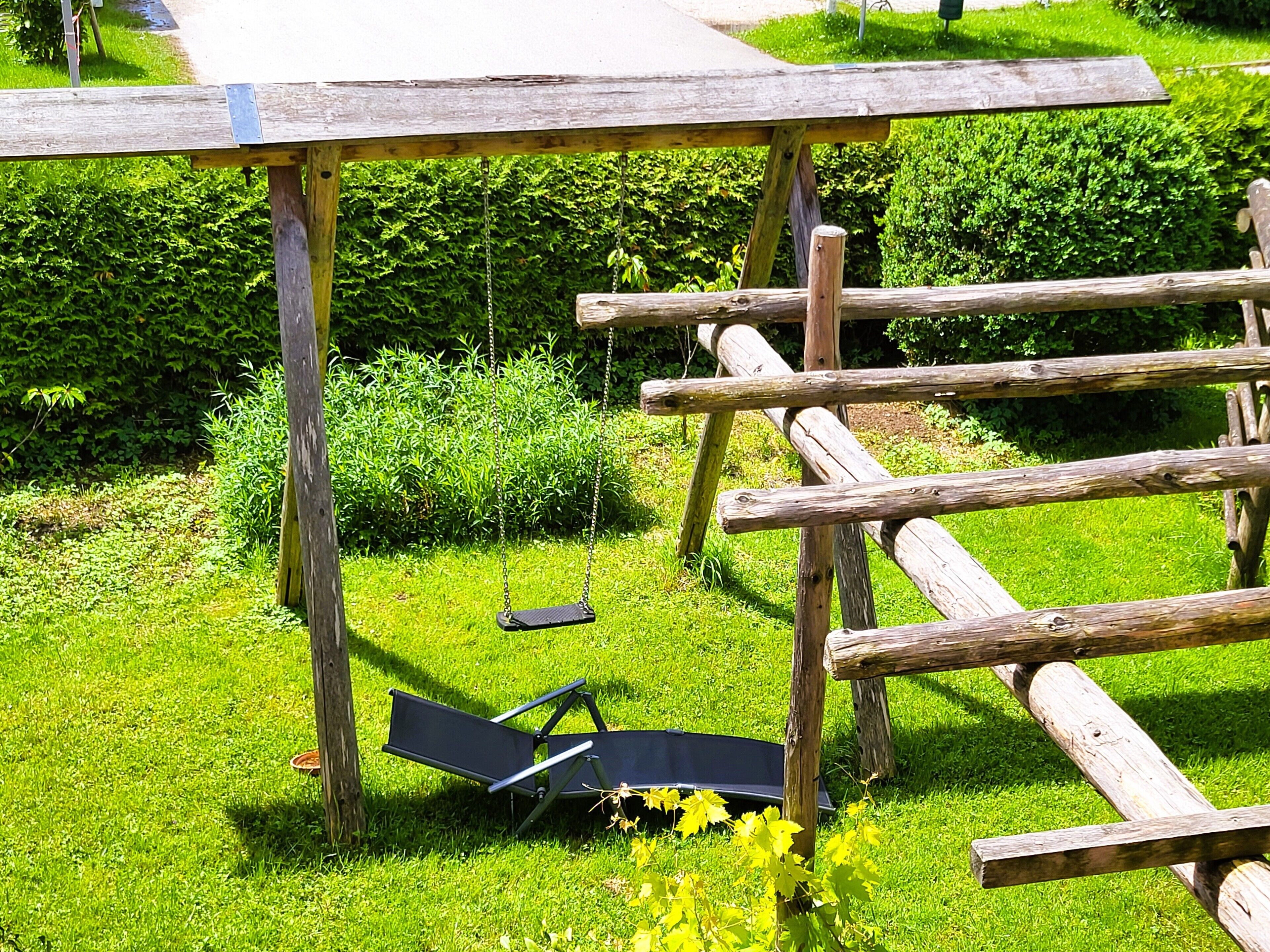 Un columpio de madera con estructura metálica y asiento negro, situado en un césped con un seto al fondo.