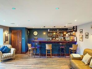 Zona de bar con mostrador de madera, taburetes y una variedad de botellas y vasos.