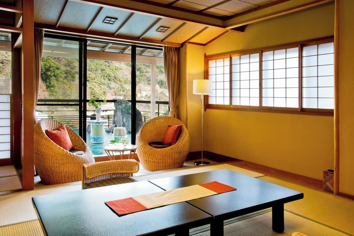A traditional Japanese room with tatami flooring, a low table, two wicker chairs, and a large window with shoji screens.