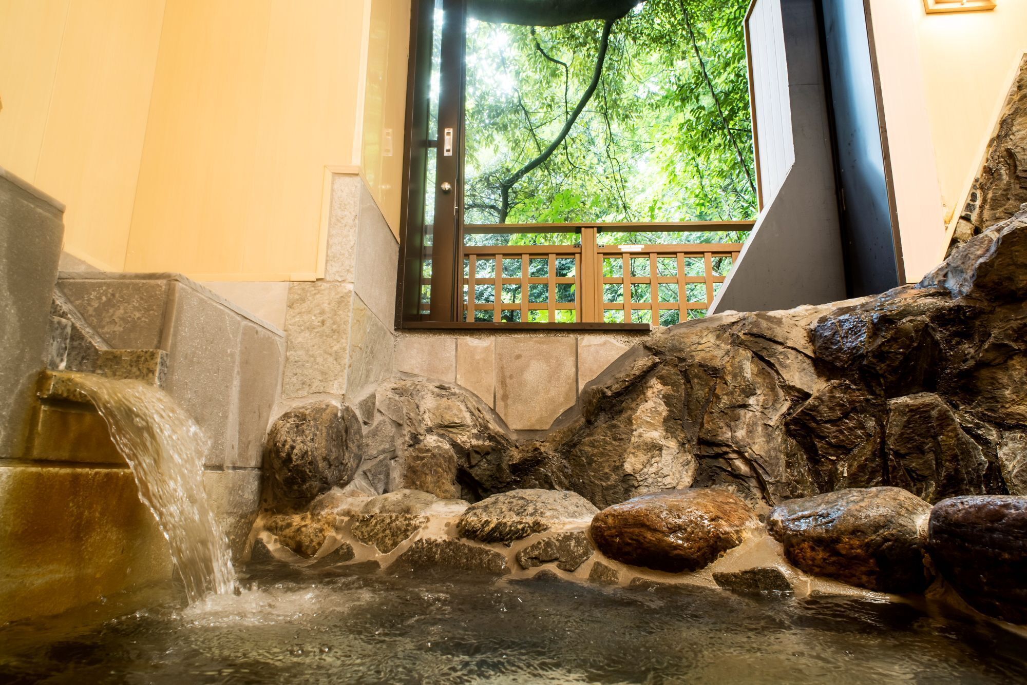 A stone water feature with a waterfall, a wooden fence, and a window with a view of trees.