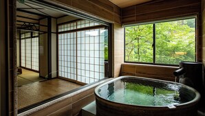 Una habitación japonesa tradicional con suelo de tatami, puerta corredera de madera, ventana grande con vistas a la vegetación y un jacuzzi de piedra.