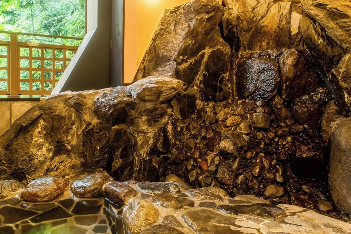 A rock formation with a water feature, a wooden fence, and a window with a view of greenery.