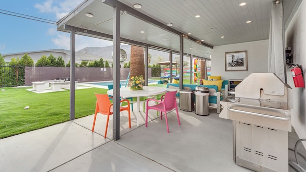 A covered outdoor patio with a grill, a table, and colorful chairs. - Palm Springs Oasis | Poolside Retreat (Palm Springs)