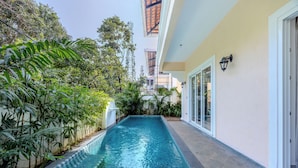 Una piscina rectangular con borde de azulejos, rodeada de exuberante vegetación y un edificio de fachada clara con un balcón que tiene techo de madera.